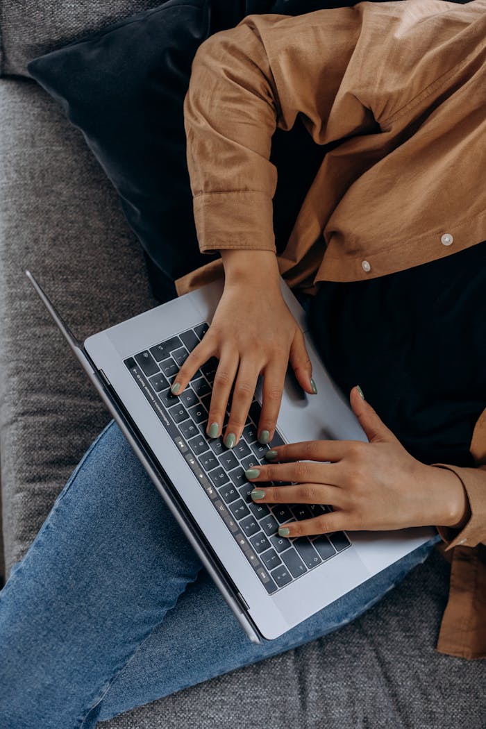 A person typing on a laptop while seated on a couch with casual attire, perfect for remote work imagery.